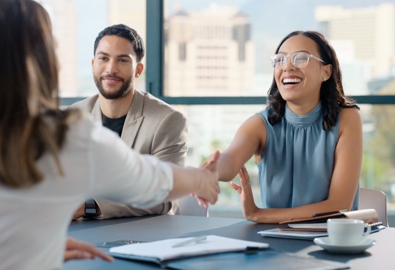 Menschen an einem Bürotisch, die sich lächelnd die Hände reichen